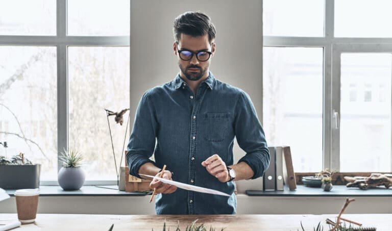 Un homme à lunettes se tient à un bureau avec une plante devant lui, s'adonnant à un vrai métier rentable.