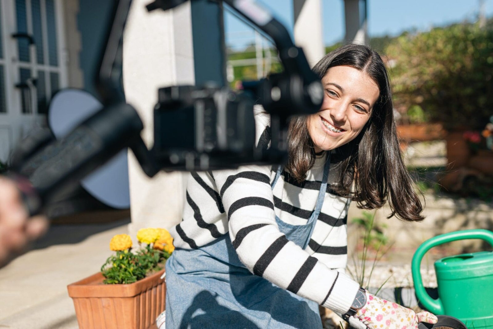 Une femme tient un appareil photo tout en arrosant ses plantes.