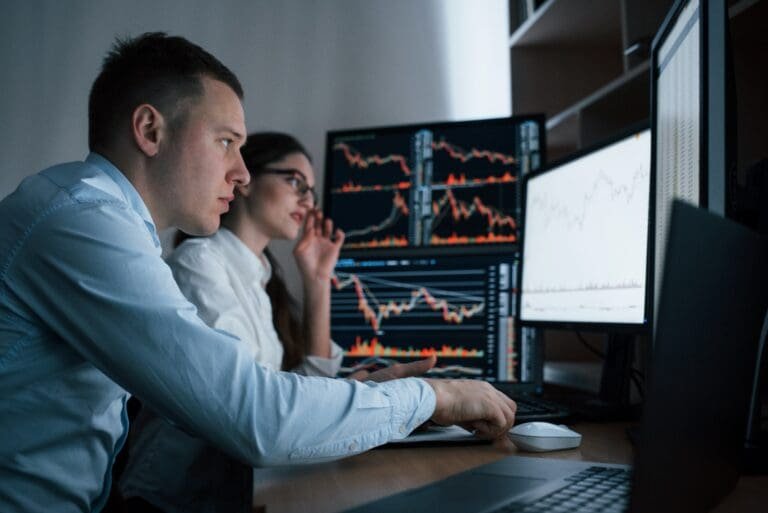 Deux personnes devant deux moniteurs avec des graphiques boursiers.